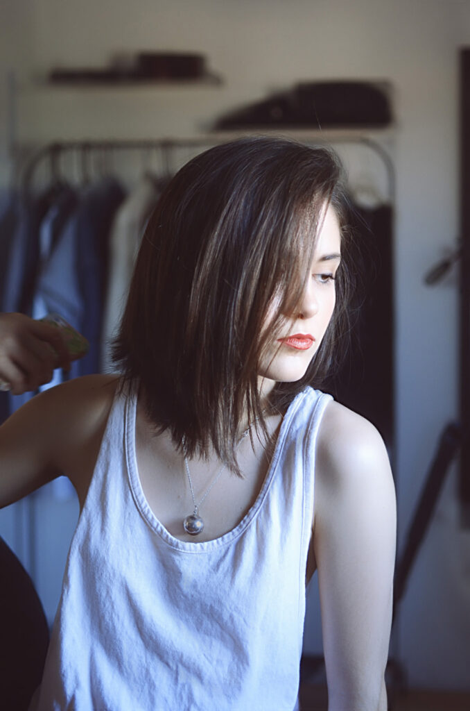 Mariana Esquivel a model with a chic bob haircut is captured in a candid moment wearing a white tank top and a silver necklace. The background reveals a room with a rack of clothes and a tripod stand.
